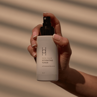 A hand holds a bottle of Skin Calm Hydrating Toner with botanical aloe extract, set against a beige background with diagonal shadow lines.