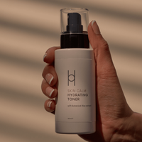 A hand holds a white bottle labeled Skin Calm Hydrating Toner, featuring natural botanical Aloe extract, set against a background with soft diagonal shadows.