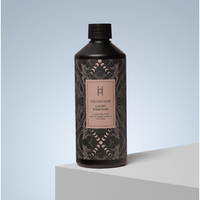 A 500ml black bottle labeled "Luxury Foam Soak - Orchid Noir" rests on a white surface with a light blue background, its elegant design suggesting soothing amber notes.