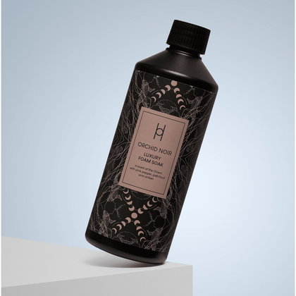 A 500ml bottle of Luxury Foam Soak - Orchid Noir, featuring a subtle floral design and infused with pink pepper, stands at an angle on a white surface against a light blue background.