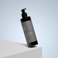 A black 250ml bottle of Moisture Protect Conditioner with pomegranate seed and baobab oils, featuring a pump dispenser and minimalist gray-toned label, is displayed on a white platform against a light gray background.