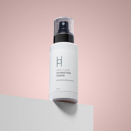 A white bottle of Skin Calm Hydrating Toner with a black spray pump sits on a white surface against a soft pink background. Its label features botanical aloe extract, making it a gentle toner for acne-prone skin.