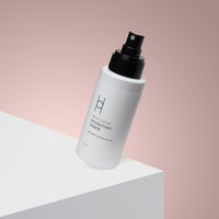 A white bottle of Skin Calm Hydrating Toner with a black spray cap is shown angled on a white surface against a soft pink background.